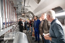 Eine Gruppe von Menschen steht in einem technischen Raum mit silbernen Rohren und Anlagen. Ein Mann in Anzug spricht, während die anderen, die in einem Mix aus formeller und legerer Kleidung sind, aufmerksam zuhören. Einige schauen zur Decke, andere direkt auf den Redner. Der Raum hat sichtbare elektrische Leitungen und Rohre, die zur Decke führen. Der Boden ist aus Gitterrosten.