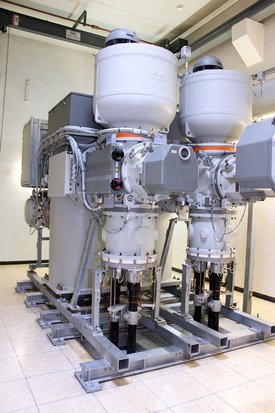 Eine technische Anordnung von zwei großen, vertikalen Maschinen, die auf einer stabilen Metallstruktur montiert sind. Jede Maschine hat einen runden oberen Teil mit einem Deckel und ist mit verschiedenen Kabeln und Rohren verbunden. An den Seiten befinden sich Bedienelemente mit bunten Anzeigen. Der Boden ist mit Fliesen ausgelegt, und die Maschinen befinden sich in einem Raum mit neutralen Wänden und Decke.