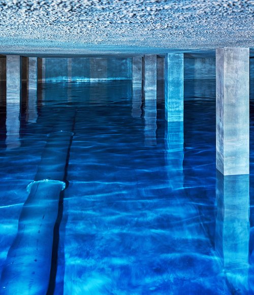 Innenansicht des Trinkwasserbehälters auf dem Sand mit spiegelglatter Wasseroberfläche, Betonstützen und indirektem blauen Licht.
