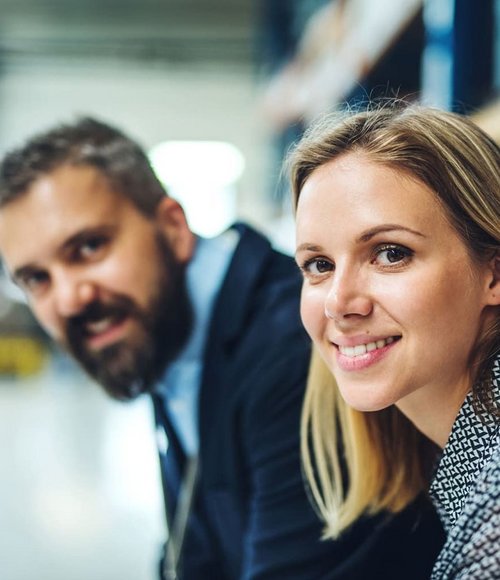 Eine Frau mit blonden langen Haaren sitzt lächelnd in einem Lagerhaus. Neben ihr ist ein Mann mit einem Bart, der ebenfalls lächelt. Im Hintergrund sind Regale und Teile eines Lagerhauses zu sehen.