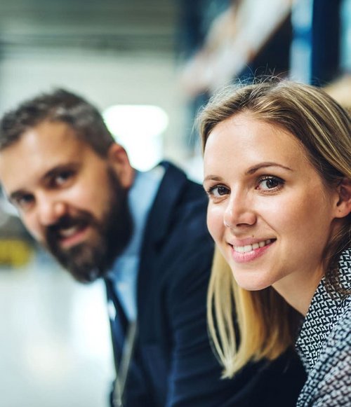 Eine Frau mit blonden langen Haaren sitzt lächelnd in einem Lagerhaus. Neben ihr ist ein Mann mit einem Bart, der ebenfalls lächelt. Im Hintergrund sind Regale und Teile eines Lagerhauses zu sehen.
